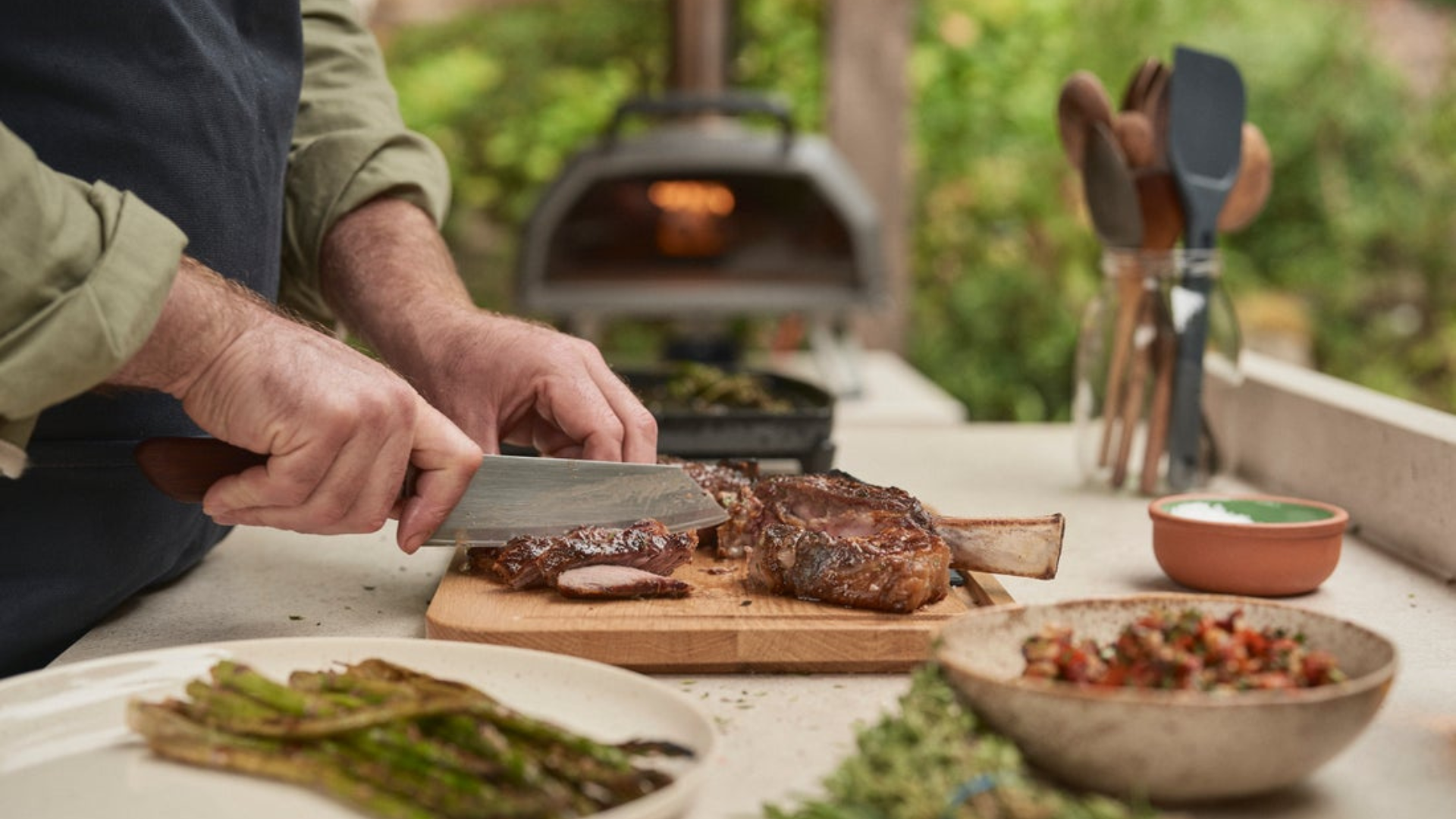 Tomahawk Steak auf Holzbrett tranchieren mit Spargel und Salsa – Ooni Karu 2 Pro als Outdoor-Küche im Hintergrund