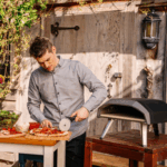Mann schneidet frische Pizza neben Ooni Koda 12 Gas-Pizzaofen im Garten – kompakter Outdoor-Pizzaofen für knusprige Steinofenpizza