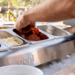 Hand greift Salamischeiben aus der Ooni Pizzabelag-Station im Freien – offene GN-Behälter mit Salami und geriebenem Parmesan auf der Ooni Topping-Station beim Outdoor-Pizzabacken