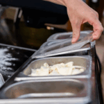 Hand hebt transparenten Deckel von Ooni GN-Behälter mit frischem Mozzarella – Nahaufnahme der Ooni Pizzabelag-Station beim Befüllen der Pizza mit Käse