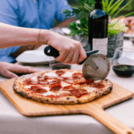 Hand schneidet Peperoni Pizza mit dem Ooni Profi-Pizzaroller auf einem Holz-Pizzabrett – geselliges Abendessen mit Rotwein und Gästen im Hintergrund