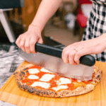 Zwei Hände schneiden eine frische Margherita Pizza mit dem Ooni Wiegemesser-Pizzaschneider auf einem Holzbrett – neapolitanischer Pizzaboden mit Tomatensoße und Mozzarella