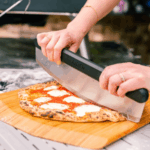 Zwei Hände schneiden eine frische Margherita Pizza mit dem Ooni Wiegemesser-Pizzaschneider auf einem Holzbrett – neapolitanischer Pizzaboden mit Tomatensoße und Mozzarella