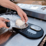 Hand legt Pizzateig-Teigling auf die große Wiegefläche der Ooni Dualen Digitalwaage – bemehlte Arbeitsfläche mit Ooni Pizzabelag-Station im Hintergrund beim professionellen Pizzabacken