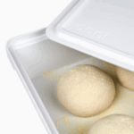 Geöffnete Ooni Pizzateigbox mit mehreren bemehlten Pizzateig-Teiglingen in der Gare – weißer Kunststoffbehälter mit Ooni-Logo-Deckel für optimale Teigruhe und Aufbewahrung