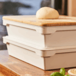 Zwei gestapelte Ooni Pizzateigboxen mit Bambusdeckel, darauf ein Pizzateig-Teigling – Nahaufnahme mit frischem Basilikum und Zitrone im Vordergrund auf Holztisch