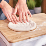 Zwei Hände formen Pizzateig auf dem Ooni Bambus-Teigdeckel und Vorbereitungsbrett – klassisches Ausdrücken des Teiglings auf dem Holzbrett mit sichtbarem Ooni-Logo
