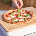 Hand legt frisches Basilikum auf fertige Margherita Pizza auf dem Ooni Bambus-Teigdeckel und Vorbereitungsbrett – perfekt gebackener neapolitanischer Pizzaboden mit Mozzarella auf Holzbrett mit Ooni-Logo
