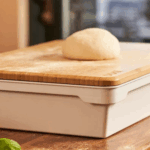 Pizzateig-Teigling auf dem Ooni Bambus-Teigdeckel, der als Deckel auf der weißen Ooni Pizzateigbox liegt – 2-in-1 Funktion als Brett und Deckel auf rustikalem Holztisch