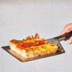 Hand hält Ooni Pizza-Servierheber mit einem Stück sizilianischer Focaccia-Pizza mit Tomatensoße und Käse – rechteckiger Pizzaschnitt auf dem Edelstahlheber beim Servieren