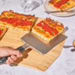 Hand hebt Stück sizilianischer Pizza mit dem Ooni Edelstahl-Servierheber vom Holzbrett – mehrere Pizzastücke mit Tomatensoße und Basilikum beim geselligen Servieren am Tisch