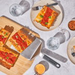 Hand hebt Stück sizilianischer Pizza mit dem Ooni Edelstahl-Servierheber vom Holzbrett – mehrere Pizzastücke mit Tomatensoße und Basilikum beim geselligen Servieren am Tisch