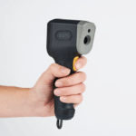 Hand hält Ooni Infrarot-Thermometer mit gelbem Auslöseknopf und Lasersensor – ergonomischer Pistolengriff des Ooni Laserthermometers zur berührungslosen Temperaturmessung am Pizzaofen