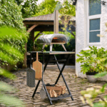 OONI Klapptisch mit Fyra 12 Holzpelletofen im Garten – Pizza-Setup mit Brennholz, Bambus- und Alu-Pizzaschieber auf Kopfsteinpflaster