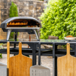 OONI Modultisch Groß mit Koda Pizzaofen und drei Bambus-Pizzaschiebern – Outdoor-Setup vor rustikaler Steinmauer