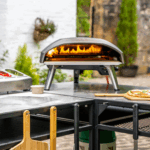 OONI Doppel-Modultisch-Setup mit Koda Pizzaofen in voller Flamme – L-förmige Outdoor-Küche mit Pizza und Zutaten vor Steinmauer