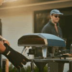 Ooni Koda 2 Pizzaofen in Dunkelblau beim Grillen im Garten mit Freunden im Hintergrund