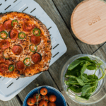 Ooni perforierter Pizzaschieber mit frisch gebackener Pepperoni-Jalapeño-Pizza, Basilikum und Kirschtomaten auf Holzuntergrund