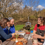 Freunde genießen selbstgemachte Pizza am Gartentisch mit dem Ooni Karu 12 Pizzaofen im Hintergrund – perfektes Outdoor-Erlebnis im Frühling