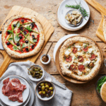 Ooni Bambus-Servierbrett mit zwei neapolitanischen Pizzen – Gemüsepizza mit Oliven und Salami-Feigen-Pizza mit Zutaten auf Holztisch