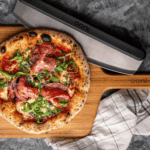 Ooni Bambus-Servierbrett mit Salami-Rucola-Pizza und Ooni Pizzaschneider aus Edelstahl auf dunklem Untergrund