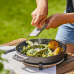 Person reibt Parmesan über gegrilltes Gemüse und Zitrone in der Ooni Gusseisenpfanne auf einem Holztisch im Garten – Outdoor-Kochen mit Ooni Pizzaofen Zubehör