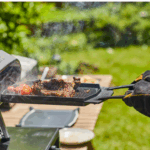 Person mit Grillhandschuhen wendet Fleisch und Tomaten auf der Ooni doppelseitigen Gusseisen-Grillpfanne – dampfendes Outdoor-Grillen direkt auf dem Ooni Pizzaofen im Garten