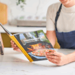 Person mit Schürze liest das Ooni Kochbuch "Cooking with Fire" in der Küche – Lifestyle-Bild des Ooni Rezeptbuchs für Pizzaofen-Rezepte und Outdoor-Kochen